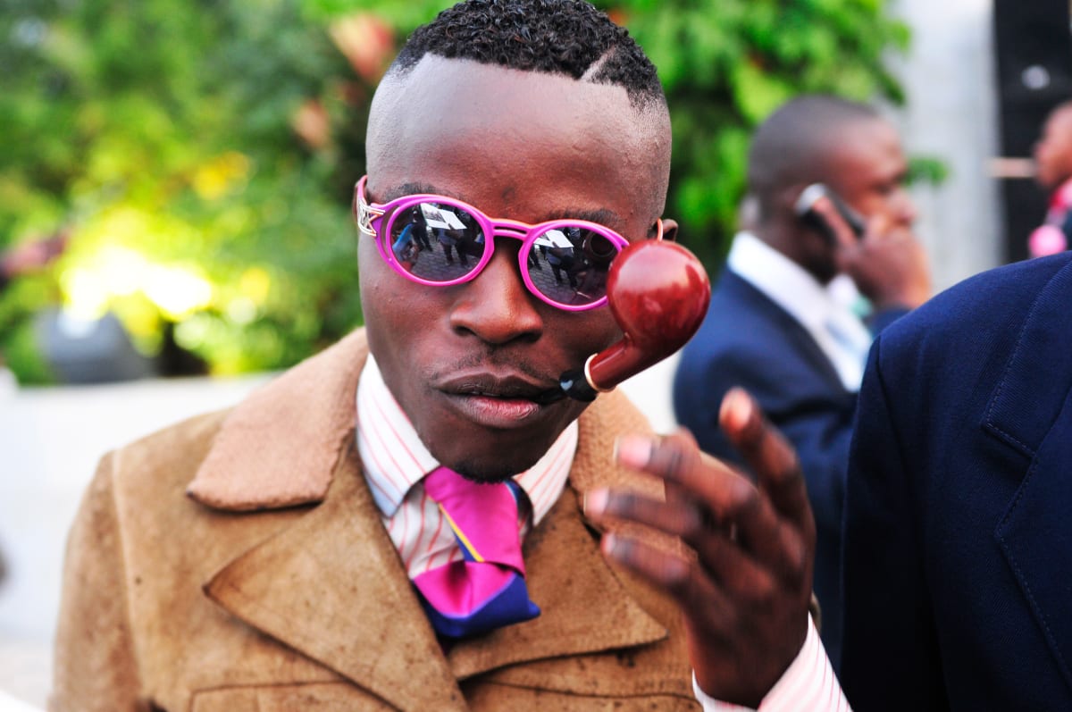 Untitled (Vive la Sape #2)  Image: Portrait of a Sapeur wearing hot pink sunglasses and matching tie, holding a pipe in his mouth. Brazzaville, Congo (2008)