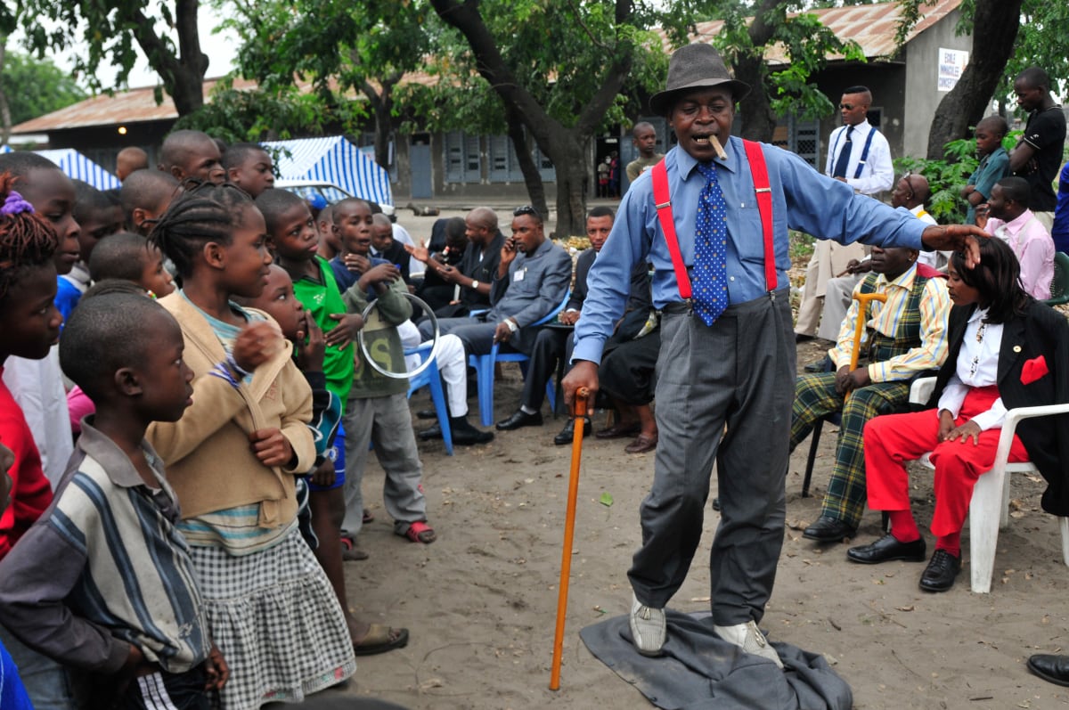 Untitled  Image: Sapeur performing in front of a crowd of kids, walking on the jacket of his suit. His look is topped by a cigar held in his lips, red bracers, a blue tie and a walking cane. Brazzaville, Congo (2008)