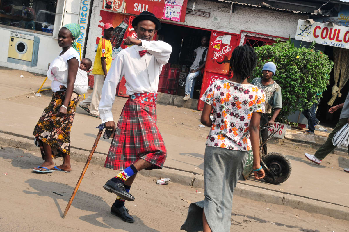 Untitled (Vive la Sape #6)  Image: Sapeur in red kilt skirt posing for the camera on the street, wearing a black bowler hat, red bowtie, white shirt, black leather shoes, walking stick and blue socks. Brazzaville, Congo (2008)