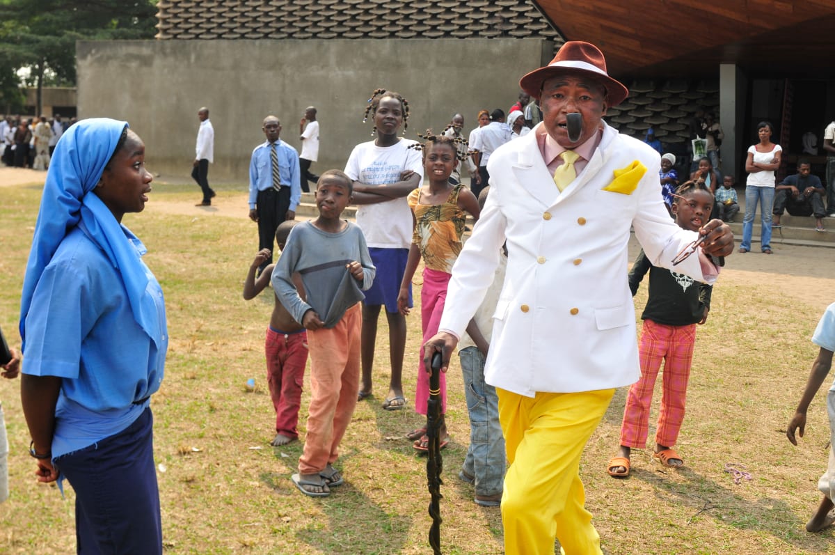 Untitled  Image: Sapeur walking for the camera wearing yellow trousers, matching pocket handkerchief, white jacket and brown hat. He is holding a pipe between his teeth and a cane in his right hand. On his left, a young nun dressed in blue is looking at him. Brazzaville, Congo (2008)