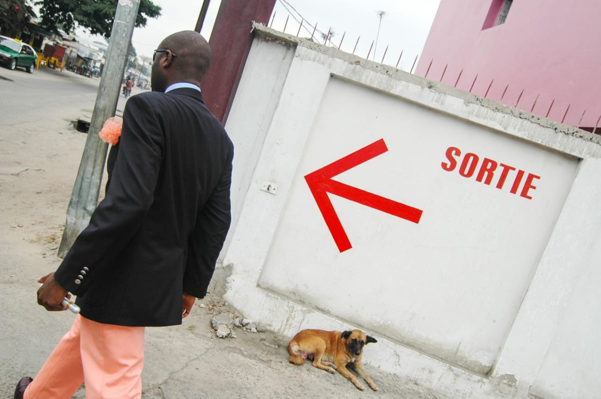 Untitled (Sortie)  Image: Salvador Hassan walking in a street of Brazzaville, with red arrow on the wall and the writing 'sortie' (exit) next to it. Salvador is the former leader of the group 'Piccadilly', one of the most innovative groups in the Sape landscape. He works for MTN, a mobile company,and as 'vestimentaire' advisor, helping people in their purchase of suits. Brazzaville, Congo (2007)