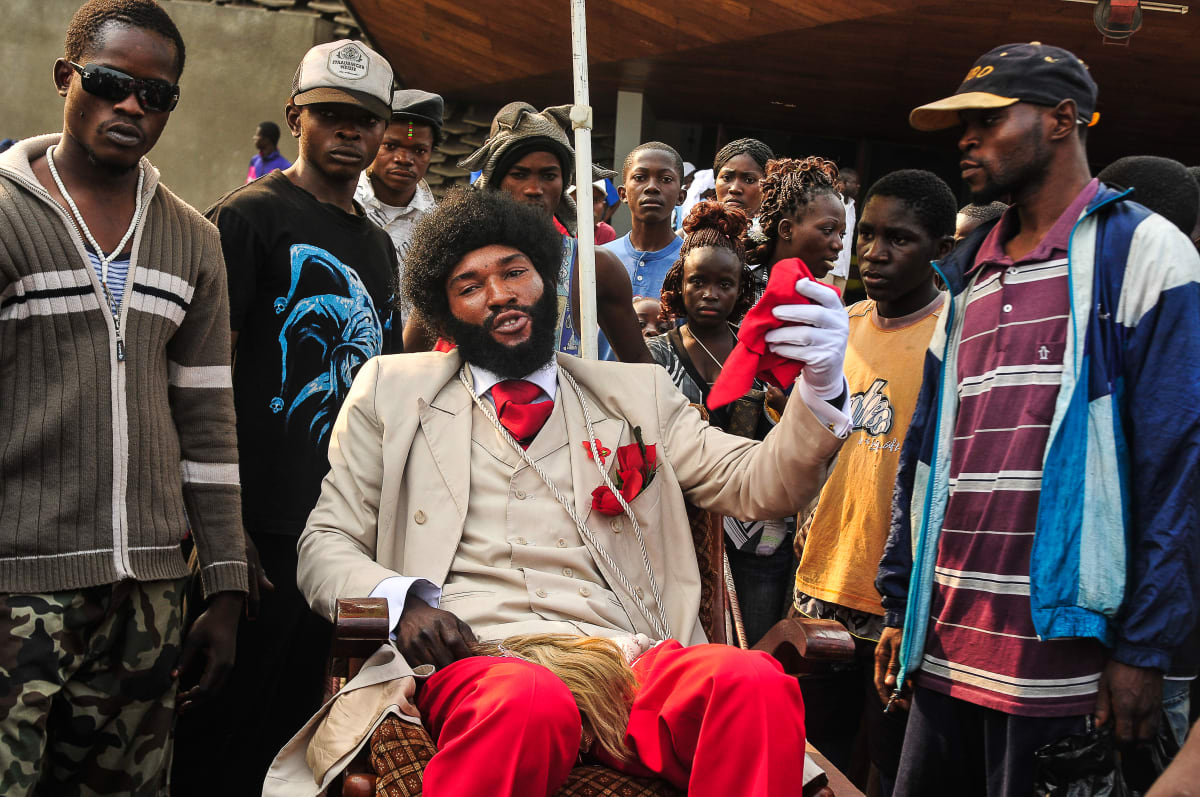 Untitled (Prince au Congo)  Image: The 'Prince of Congo' sitting on his charriot-throne, surrounded by a crowd. He is wearing red trousers and matching tie and pocket handkerchief, beije jacket and white gloves. Brazzaville, Congo (2008)