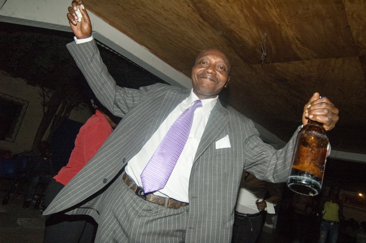 Untitled  Image: Man at a club in grey pinstripe suit and lilac cravat with his arms held high and a bottle in the left hand. Brazzaville, Congo (2007)
