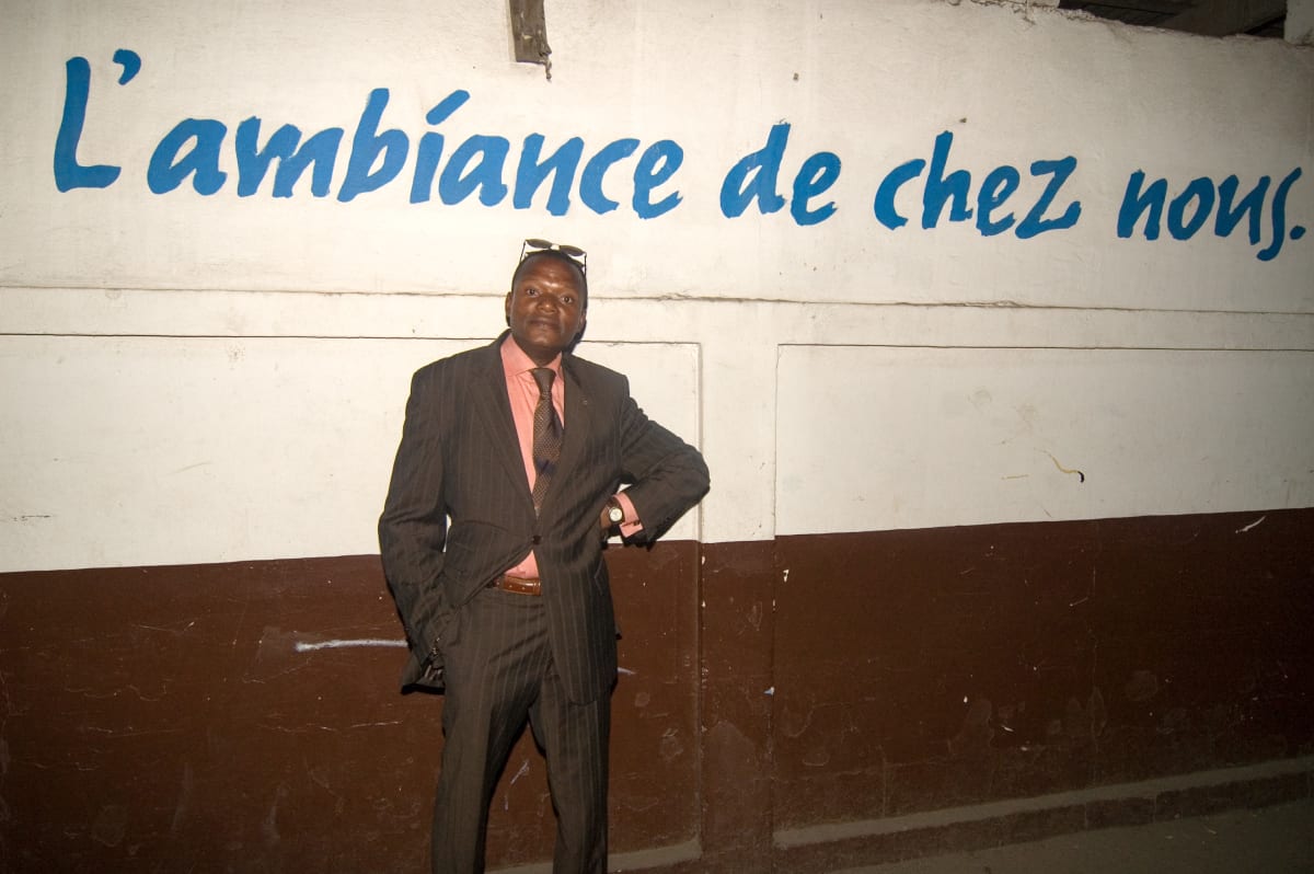 Untitled  Image: Night scene of a man in brown pinstriped suit and pink shirt standing before a wall with the writing 'L'ambiance de chez nous' (The atmosphere of home). Brazzaville, Congo (2007)