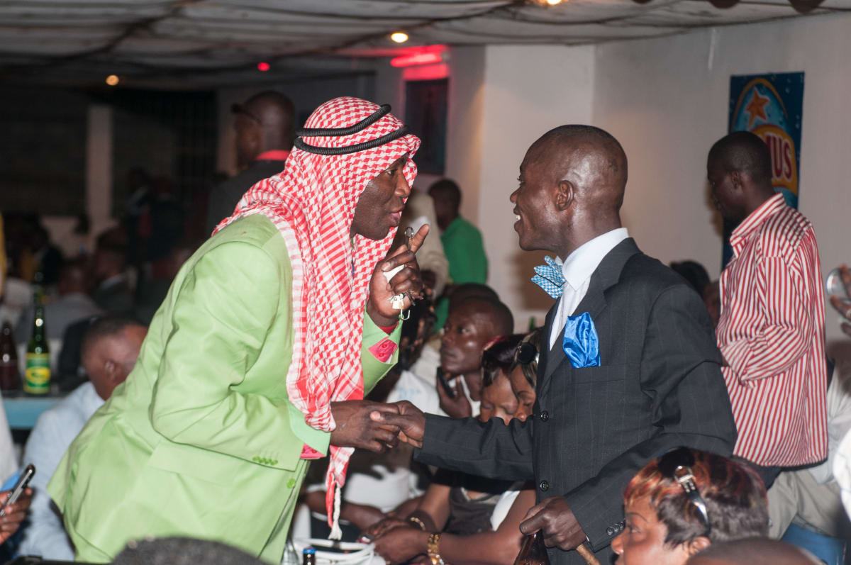 Untitled  Image: Man with a red kefia on his head and bright green jacket shaking hands and talking with a man in dark blue pinstriped suit in a nightclub. Brazzaville, Congo (2008)