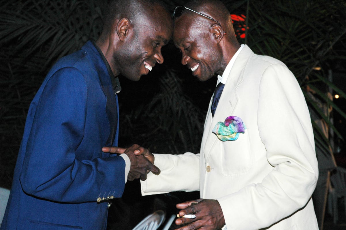 Untitled  Image: Two men in a club, one in blue suit, the other in white suit, saluting each other by leaning forehead to forehead and shaking their hands. Brazzaville, Congo (2007)