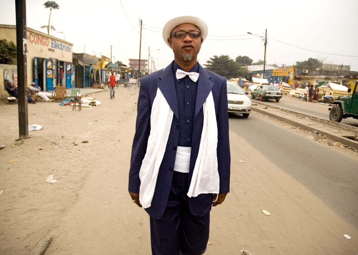 Untitled (Kvv Mouzieto in Avenue Matsoua) by Daniele Tamagni Foundation  Image: Jeanclaude, who goes by the name of 'Kvv Mouzieto', walking down Avenue Matsoua. He is a famous Sapeur of Brazzaville, and belongs to the second generation of Sapeurs. He is wearing a blue suit with matching shirt, a white bowtie and matching scarf, topped by a white hat. Brazzaville, Congo (2007).