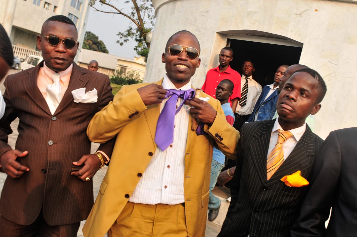 Untitled  Image: Group of three sapeurs at a party. The man in the center, in a yellow suit, is untying his tie. Brazzaville, Congo (2008)