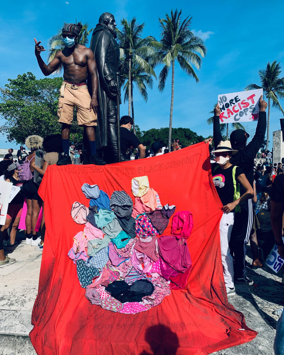 Arpillera Americanx * Cunt Quilt (Crown) Cunt Congress by Coralina Rodriguez Meyer  Image: A fellow protester drapes Arpillera Americanx Cunt Quilt (Crown) on the Colonizer statue's feet while standing on his pediment during the George Floyd protest May 31, 2019 in downtown Miami