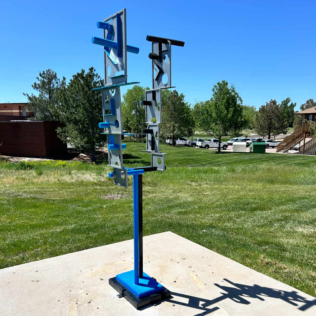 Split Decision (Art Encounters, 2024) by Andrew Libertone  Image: Colorado artist Andy Libertone’s architecturally modern sculpture, “Split Decision,” sits prominently at the corner of Mainstreet and E. Mainstreet. Andy is well-known to the local arts scene, having taught at many local institutions. He also is a founding member of Spark co-op gallery and has exhibited his work in galleries, institutions, and the public throughout Colorado and the west. The motivation for his geometric sculptures, such as this one, comes from a combination of his love of jazz music, architecture, industrial and, auto-based imagery. His large geometric, powder-coated steel sculptures demonstrate his artistic balance of solid forms, open spaces, and orchestrating colors in steel and wood—a challenge that’s proven to be a rewarding expansion of his artistic vision.