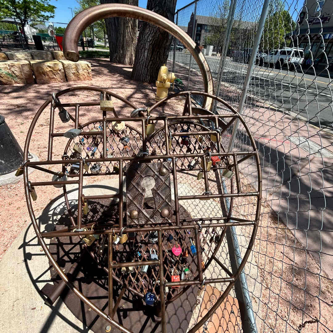 Unconditional (Art Encounters, 2024) by Mitch Levin  Image: Boulder artist Mitch Levin uses found metals whose patina tells the story of time. He especially enjoys the connectivity of humans and this piece is no exception, inspired by his visit to Paris, where he says the lock bridge there was the catalyst to this work. “I felt a traveling piece like this would leave its mark on the audience and in return they would indelibly leave their mark on the piece, as well as being part of the art process.” In fact, he hopes those who visit his sculpture will attach their own personal locks, thus connecting them in the aesthetics of his work. “Unconditional” is located at the Southeast corner of O’Brien Park.

