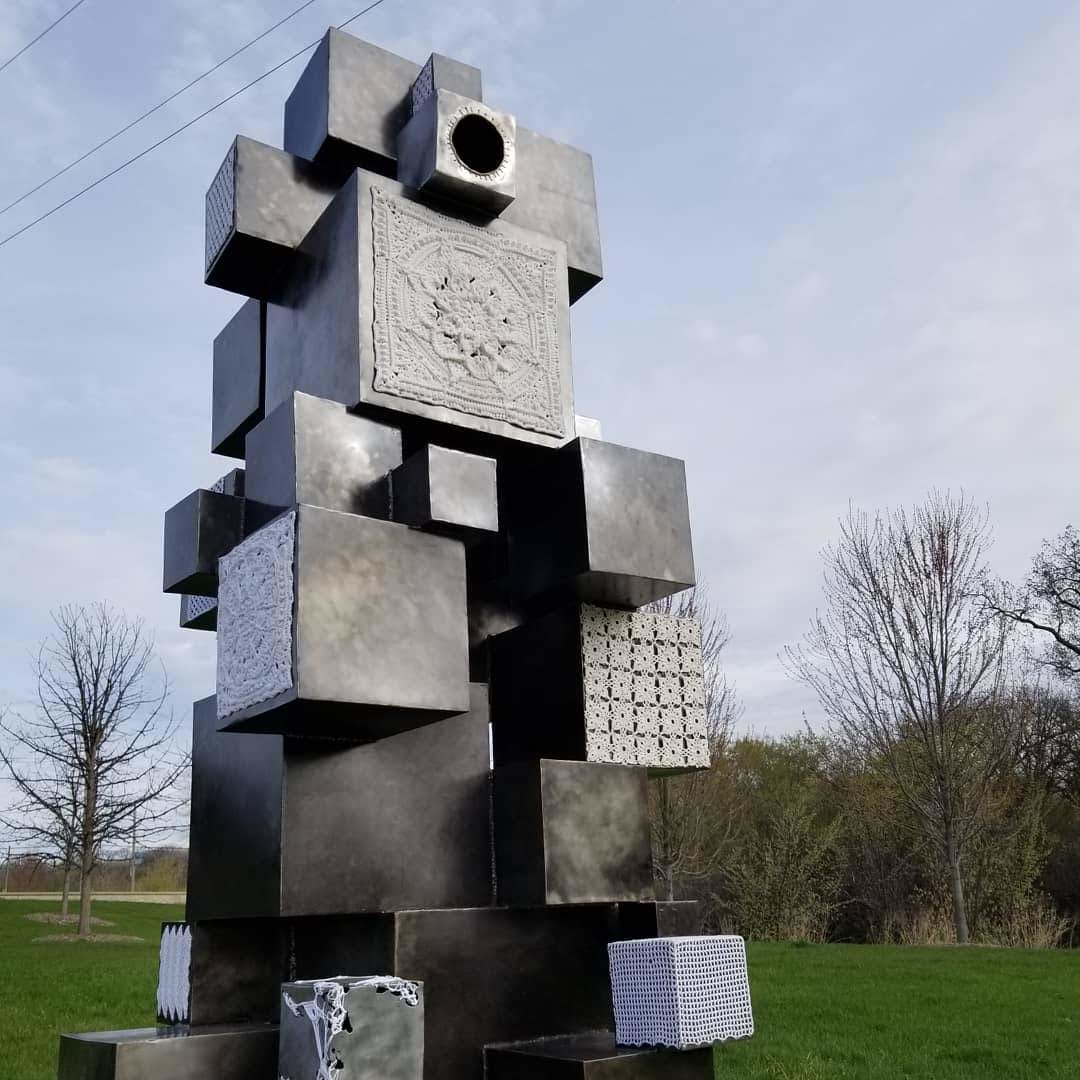Elaborated by Amanda Langer  Image: 8 foot tall sculpture of haphazardly stacked steel cubes of various sized, welded together.  Several cube surfaces covered with crocheted fiber patterns.