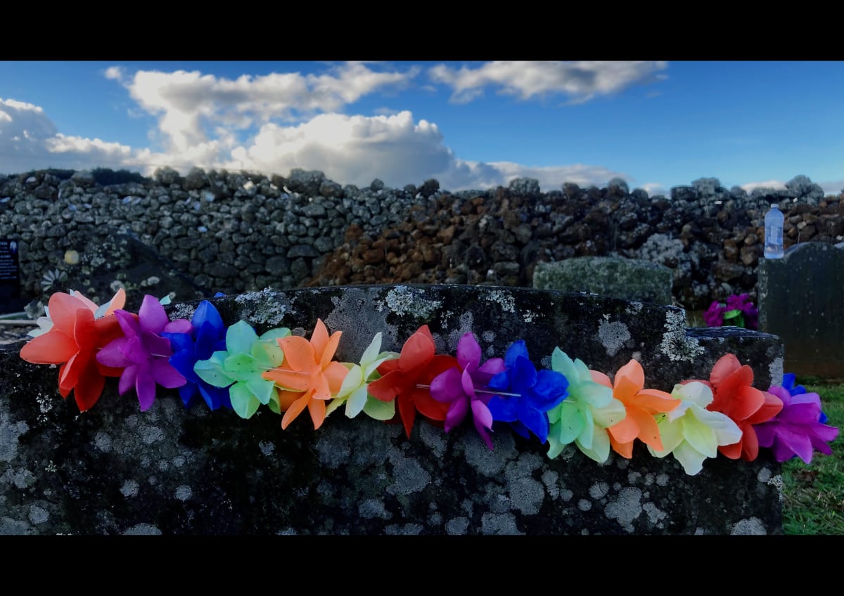 Lei over gravestone, rock wall and sky, St Michael's urupā, Ōhaeawai, 30 April 2021 by Dr  Rangihiroa Panoho  Image: This the site of the original battle at Pene Taui's pā redesigned by Te Ruki Kawiti at Ohāeawai. As at Ruapekapeka the fortification caused the British enormous embarrassment and was not taken destroyed until Kawiti, Heke and their men had deserted the trenches. The church and cemetery sit atop the original battle site which historically has become a way of de-commissioning a site of conflict.
