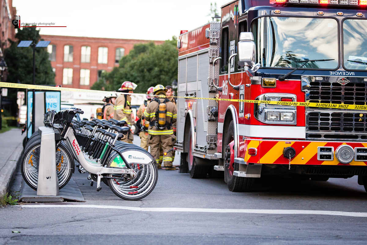 Zone Urgence by André JAY Chapdelaine  Image: Emergency Response: A scene of urgency and calm, where the vibrant red of a firetruck contrasts with the stillness of parked bicycles. This photograph captures the duality of urban life—action and pause, chaos and order—through a moment in Montreal’s