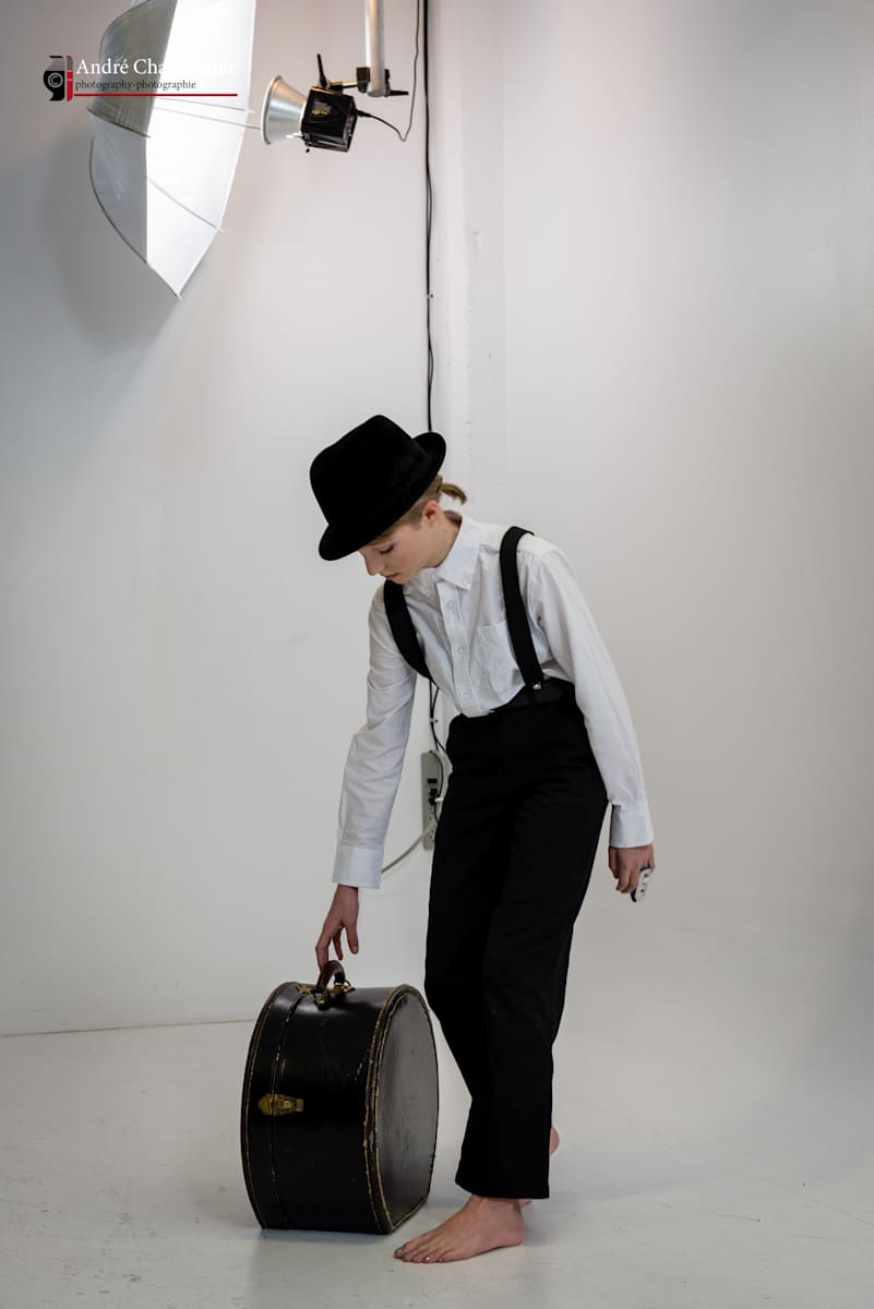 Taking a Tryp by André JAY Chapdelaine  Image: Une prise de vue en studio minimaliste d’un jeune voyageur en tenue vintage, tenant une valise. Faisant partie de la série « Les Anges d’André », l’image saisit un moment de calme anticipation et le début d’un voyage. 
A minimalist studio shot of a young traveler, dressed in vintage attire, holding a suitcase. Part of the "Andre's Angels" series, the image captures a moment of quiet anticipation and the beginning of a journey