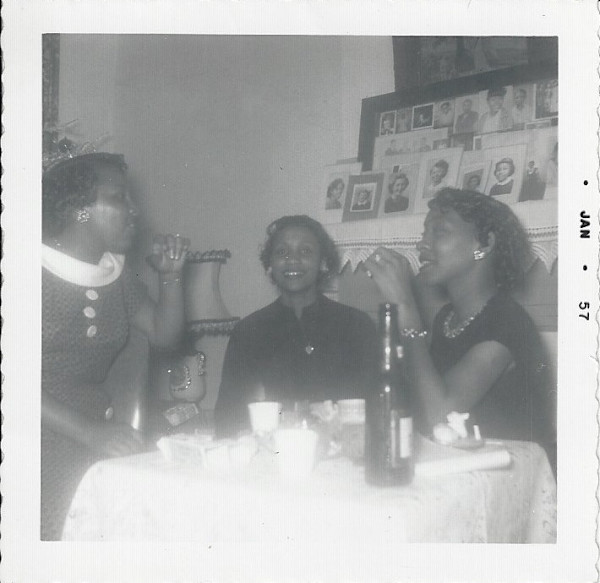Three women at a table with photographs on mantle by Unknown
