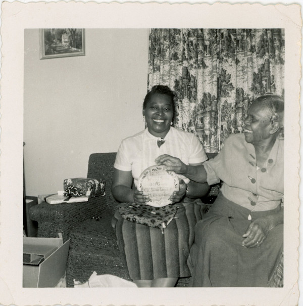 Woman accepting kitchen plaque gift from another woman by Unknown