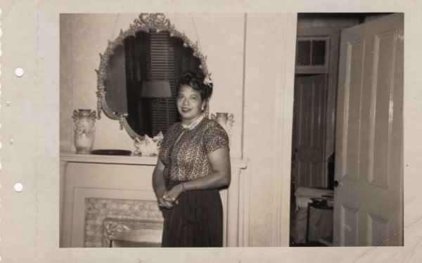 Woman standing by fireplace in front of mirror by Unknown