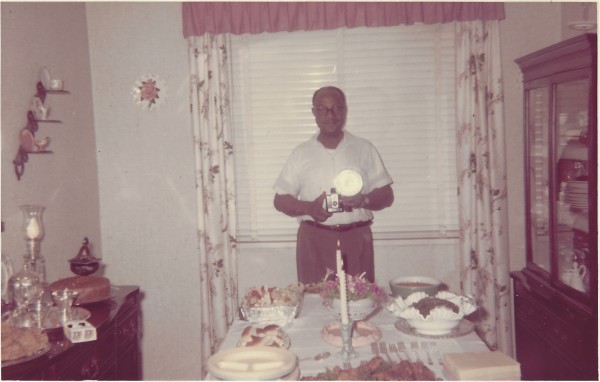 Man standing at dining table holding Brownie camera with flash by Unknown