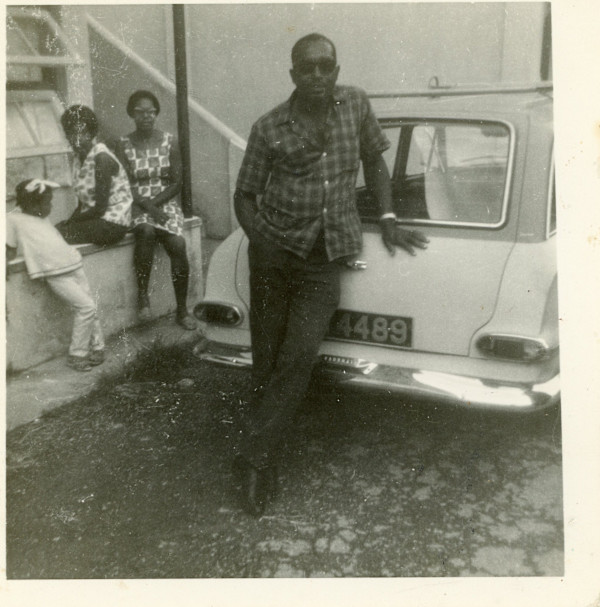 Man leaning on car; women sitting nearby by Unknown