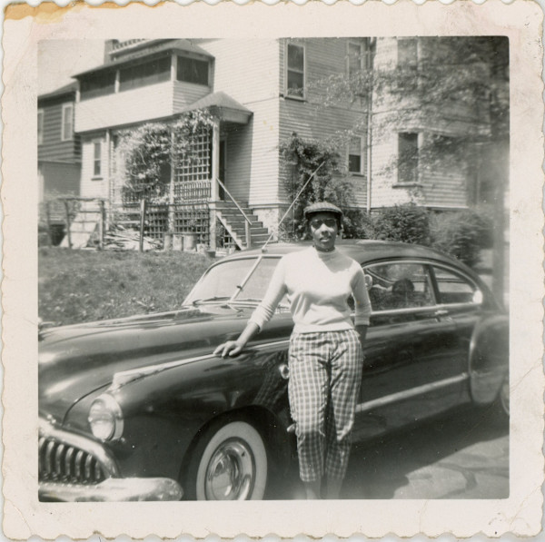 Woman wearing checkered pedal pushers and kangol hat standing next to automobile by Unknown