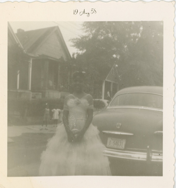 Girl in formal dress standing behind automobile by Unknown