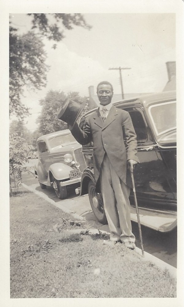 Man standing next to automobile in top hat and tails by Unknown