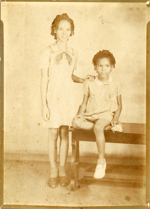 One girl standing, one girl sitting on bench by Unknown