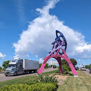 A commercial semi truck drives by a tall, abstract sculpture on a green median.