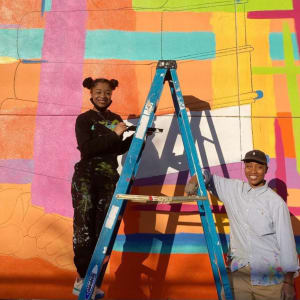 Colors of Our Culture by Young Artist Movement (YAM), Journey Allen  Image: YAM artist Aliyah Pflueger with mentor Journey Allen as the two work on installing a mural on New Orleans East Regional Library's Read Boulevard wall 